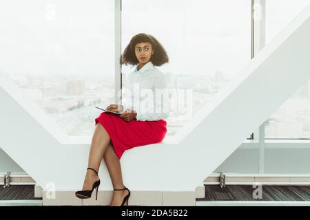Junge hinreißende schlanke afroamerikanische Geschäftsfrau sitzt auf einem Zeitgenossen Fensterbank des modernen Büros mit einem digitalen Tablet In Händen A Stockfoto