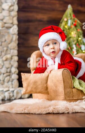 Nahaufnahme Porträt eines kleinen Jungen als roter Weihnachtsmann gekleidet, spielen am Steinkamin mit hellen Girlanden, setzen eine Geschenkbox unter Weihnachtsbaum Stockfoto