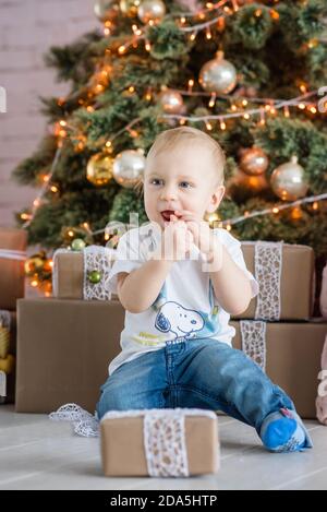 Kleiner blonder Junge, der einen Lebkuchenmann am Weihnachtsbaum zu Hause isst. Neujahrskarte mit Girlanden Bokeh. Das Baby lacht, öffnet eine Geschenkbox Stockfoto