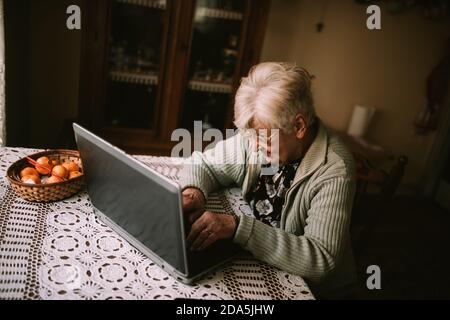 Ältere Großmutter sitzt am Tisch und tippt Nachrichten an Laptop zu ihrer Familie während Quarantäne COVID - 19 Coronavirus Stockfoto