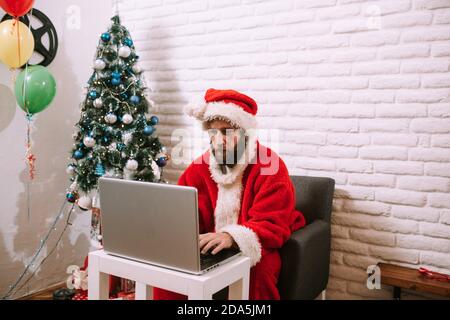 Der Weihnachtsmann sitzt am Tisch mit einem Laptop neben dem Weihnachtsbaum bei ihm zu Hause. Weihnachtszeit. Stockfoto