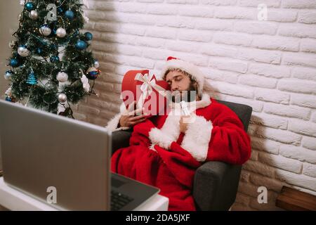 Der Weihnachtsmann schläft auf einem Sessel neben einer roten Geschenkbox, vor einem Tisch mit Laptop. Weihnachtsmann feiert Weihnachten und Neujahr online Stockfoto