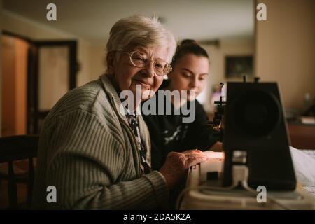 Eine süße alte kaukasische Großmutter lehrt ihre Enkelin, an einer Nähmaschine zu nähen. Oma schaut auf die Kamera Stockfoto