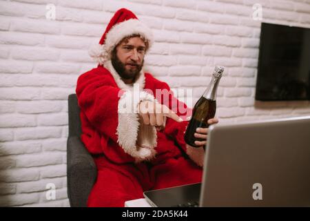 Der Weihnachtsmann sitzt im Haus und zeigt auf den Laptop, während er eine Flasche Champagner hält. Santa Claus online feiert Neujahr Stockfoto