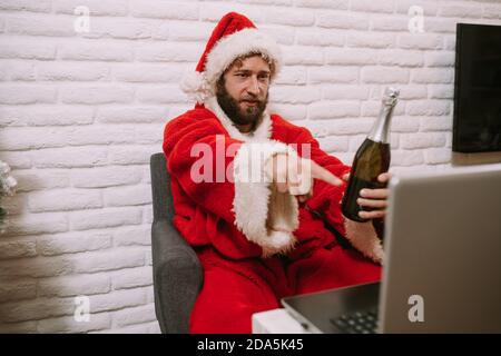 Ein kaukasischer Mann in einem Weihnachtsmann-Anzug sitzt im Haus mit einer Flasche Champagner in der Hand und spricht über einen Videoanruf über einen Laptop. Stockfoto