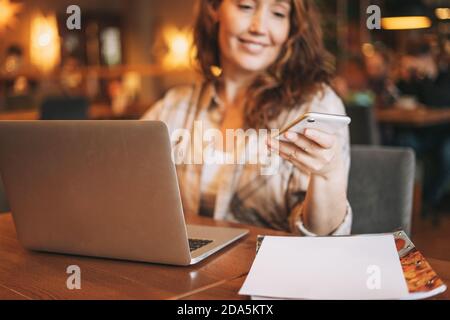 Erwachsene charmante Brünette Frau in karierten Hemd arbeiten mit Laptop mit Handy im Café, konzentrieren sich auf die Hand Stockfoto