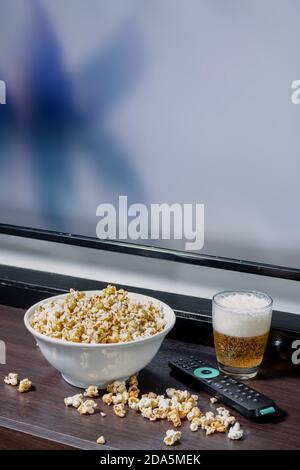 Eine Schüssel voll Popcorn, etwas Popcorn fiel auf den Tisch neben einem Lager vor einem LED-fernseher mit Fernbedienung. Stockfoto