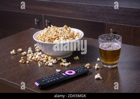Eine Schüssel voll Popcorn, etwas Popcorn fiel auf den Couchtisch neben einem Lager vor einem LED-fernseher mit Fernbedienung. Stockfoto