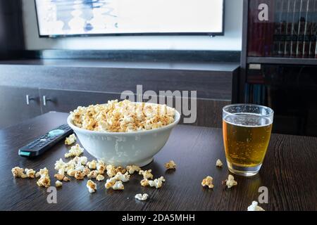 Eine Schüssel voll Popcorn, etwas Popcorn fiel auf den Couchtisch neben einem Lager vor einem LED-fernseher mit Fernbedienung. Stockfoto