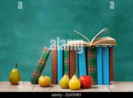 Offenes Lehrbuch, Stapel von Büchern in bunten Abdeckungen, Birnen und Äpfel auf Holztisch mit grünem Tafelhintergrund. Fernunterricht. Zurück zu s Stockfoto