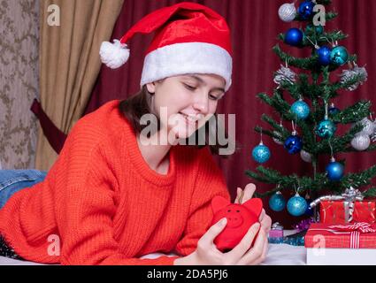 Lächelndes Teenager-Mädchen in einem Weihnachtshut und roten Pullover nimmt Münzen von einem roten Sparschwein zum Einkaufen Weihnachtsgeschenke. Online-Shopping-Konzept, c Stockfoto