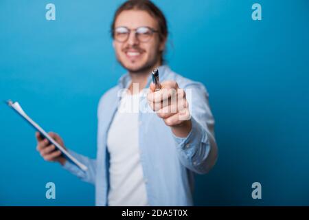 Junger bärtiger Geschäftsmann mit Brille und langen Haaren hält Einige Dokumente und zeigen mit dem Stift auf die Kamera lächelnd zahnhaft Stockfoto