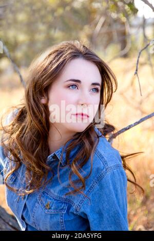 Schöne junge Frau mit goldbraunem Haar, blauen Augen und heller Haut in blauem Denim-Kleid, das ruhig vom Betrachter weg aussieht Stockfoto