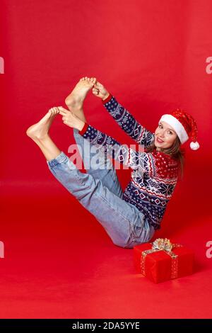 Eine in voller Länge lächelnde Frau in einem Weihnachtspullover und Weihnachtsmütze spielt Sport, isoliert auf einem roten Hintergrund Stockfoto