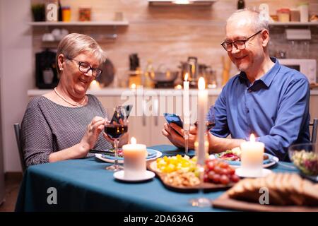 Pensionierter Mann, der auf dem Smartphone in der Küche browsen und mit seiner Frau zu Abend essen kann. Sitzen am Tisch in der Küche, Surfen, Suchen, Telefon, Internet, feiern ihr Jubiläum im Speisesaal. Stockfoto