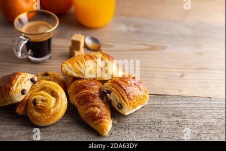 Frühstück mit Croissants, Zimtbrötchen und Schokoladeplätzchen, frischem Orangensaft und Kaffee auf der Seite auf Holzgrund. Stockfoto