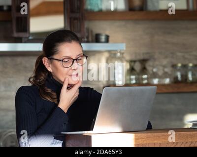 Kaukasische Geschäftsfrau mittleren Alters mit Brille, die in der Küche mit Laptop und Dokumenten arbeitet. Bleiben Sie zu Hause während covid-19 Quarantänekonzept. Stockfoto