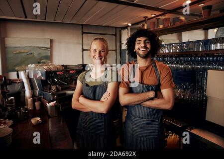 Portrait von jungen weiblichen und männlichen Mitarbeitern mit Schürze stehend Hinter dem Tresen im Café mit gekreuzten Händen und Blick auf die Kamera Stockfoto