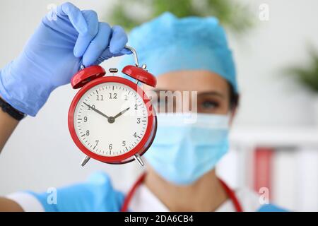 Arzt in der medizinischen Schutzmaske hält roten Wecker in Seine Hände Stockfoto
