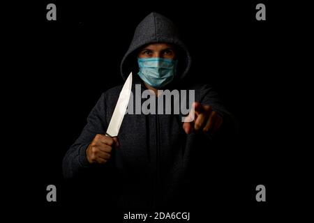 Junger Mann in medizinischer Maske zeigt Finger, während er Messer hält Auf schwarzem Hintergrund Stockfoto