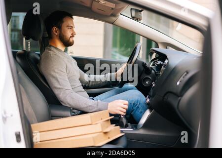 Seitenansicht des bärtigen Lieferers in legerer Kleidung, der mit dem Auto fuhr, brachte dem Kunden heiße Pizza. Stockfoto