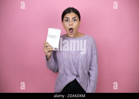 Schöne Frau trägt einen lässigen violetten Pullover auf rosa Hintergrund Erschrocken und überrascht halten Notebook ohne Wort Nachricht Stockfoto