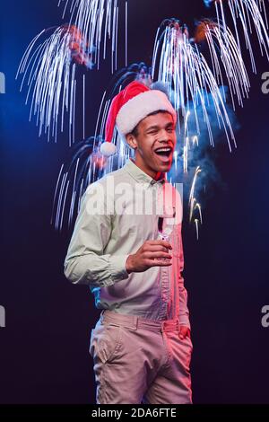 Heiraten Sie Weihnachten. Vertikale Aufnahme eines jungen aufgeregten Mixed-Race-Mannes, der die Mütze des Weihnachtsmannes mit einem Glas Champagner trägt und vor dem Hintergrund eines Feuerwerks steht. Menschen und Urlaub Stockfoto