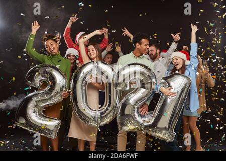 Frohes neues Jahr und heiraten Weihnachten. Junge multirassische Menschen halten silberne Folienballons in Form von Zahlen 2021 und tanzen, Konfetti fallen in die Luft. Feiertags-, Party- und Feiertagskonzept Stockfoto