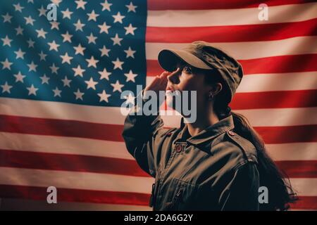 Memorial Day. Eine Soldatin in Uniform grüßt vor dem Hintergrund der amerikanischen Flagge. Seitenansicht. Das Konzept des amerikanischen Nationalfeiertags Stockfoto