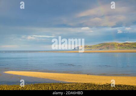 Schwacher Regenbogen über dem ruhigen blauen Meer und der Landspitze von Llanddona von Red Wharf Bay, Isle of Anglesey, Wales, Großbritannien Stockfoto