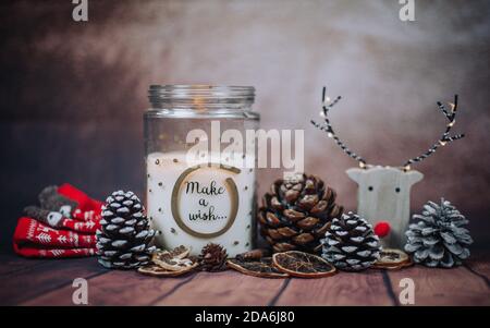 Weihnachtskomposition mit "Make a wish" Kerze in einem Glas mit Regenhirsch, Weihnachtsbeleuchtung und Tannenzapfen auf einem rustikalen braunen Hintergrund. Weihnachten Stockfoto