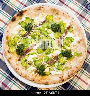 Eine echte italienische Gemüsepizza in einem Gericht, Zucchini, Sardellen und Brokkoli Blick von oben auf einer karierten Tischdecke Stockfoto