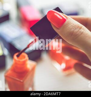 Weibliche Finger halten Pinsel mit orange Nagellack. Hintergrund ein Tisch mit einer Reihe von Nagellacken in verschiedenen Farben Stockfoto