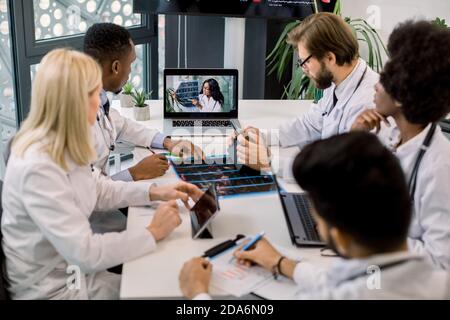 Team von multirassischen erfahrenen Ärzten oder Wissenschaftlern, mit Videokonferenz mit ihren jungen zuversichtlich afrikanischen Expertin und sprechen über die Stockfoto