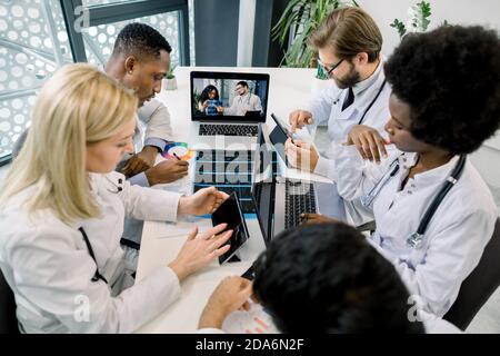 Multiethnische Ärzte machen Notizen auf Laptop und Tablets während eines Videoanrufs im Krankenhaus, im Chat mit zwei selbstbewussten afrikanischen und kaukasischen Ärzten Stockfoto