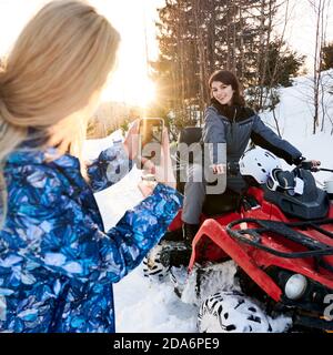 Schöne Brünette Frau sitzt auf All-Terrain-Fahrzeug und lächelt, während Freund fotografieren mit Smartphone. Junge Frau auf dem Quad am sonnigen Tag in den Winterbergen. Konzept des Quad-Bikens. Stockfoto