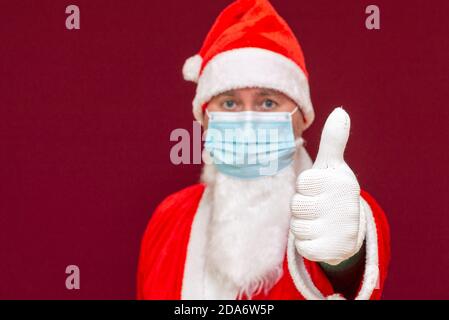 Nahaufnahme Santa Claus Tragen medizinische Maske zeigt Daumen nach oben Neujahr und frohe Weihnachten und frohe Feiertage Konzept.Red Hintergrund.Selective Fokus. Stockfoto