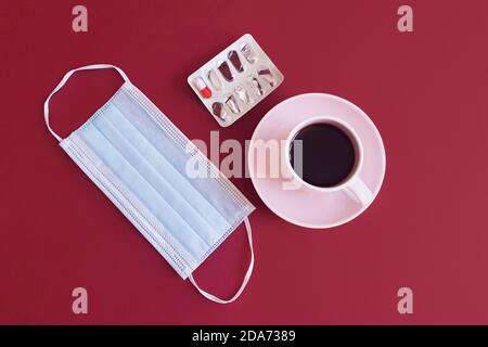 Tasse Kaffee, entfernte medizinische Maske und offene Pillenpackung auf rotem Tisch. Draufsicht. Stockfoto