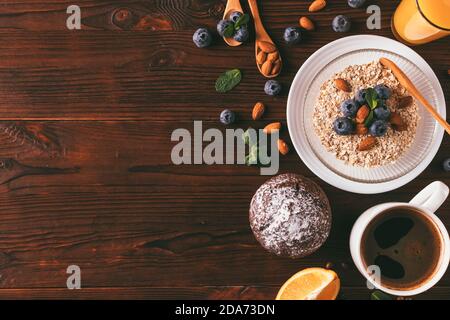 Frühstück mit Haferflocken, Mandeln und Heidelbeeren neben einer Tasse Kaffee und Schokoladenmuffin auf einem Holztisch, Blick von oben. Stockfoto