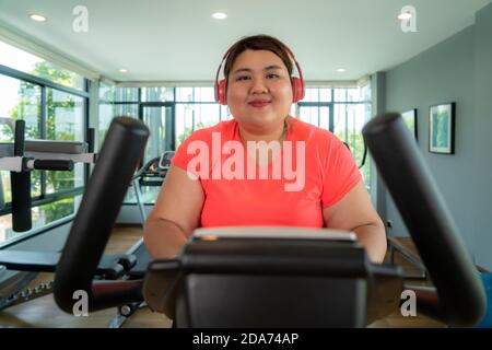 Übergewichtige asiatische Frau mit Ohrhörertraining auf dem Heimtrainer in der modernen Sporthalle, glücklich und lächelnd beim Workout. FAT Woman kümmert sich um Gesundheit und Stockfoto