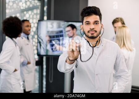 Porträt eines jungen indischen Arztes mit Stethoskop, Blick auf die Kamera, während seine multiethnischen Kollegen im Hintergrund, mit tv-Bildschirm, diskutieren Stockfoto