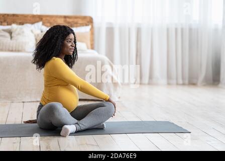 Sportliche schwarze schwangere Frau sitzt auf Yoga-Matte, Training Stockfoto