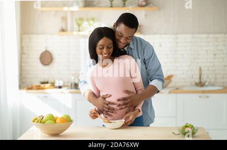 Ziemlich schwarz schwangere Frau Kochen gesundes Frühstück in der Küche und Ihr Freund umarmte sie von hinten Stockfoto