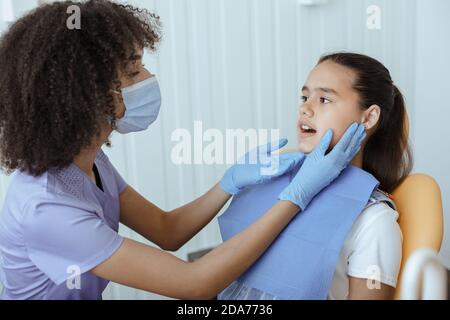 Junge ernsthafte afroamerikanische Arzt in Uniform, Schutzmaske und Handschuhe prüft Patienten Mund Stockfoto