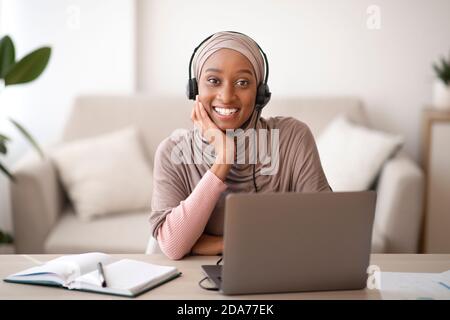 Portrait der schönen muslimischen schwarzen Frau im Kopftuch trägt Headset, online kommunizieren auf Laptop-Computer, drinnen Stockfoto