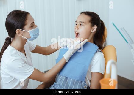 Junge männliche Arzt in weißen Uniform, Schutzmaske und Gummihandschuhe untersucht Zähne des kleinen Mädchens Stockfoto