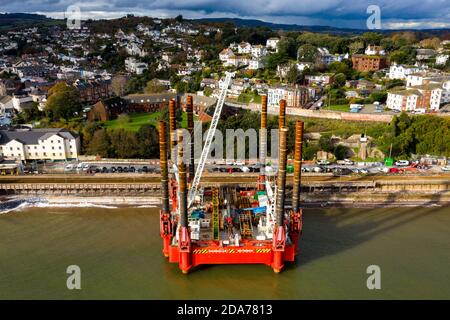 Sonnenlicht fällt auf eine riesige Baumaschine am Bahnhof Dawlish, wo mit dem Bau einer neuen und größeren Meeresmauer begonnen wurde, um die Eisenbahn und die Stadt mit einem achtbeinigen, in sich geschlossenen Wagenkahn vor steigenden Meeresspiegel zu schützen. Bekannt als Wavewalker – der einzige seiner Art in Europa. Es ist das erste Mal, dass diese Art von Lastkahn verwendet wurde, um das britische Schienennetz zu unterhalten und die Maschine ist in der Lage, über hohe Gezeitenbereiche zu fahren, die besonders die Südküste von Devon beeinflussen. Stockfoto