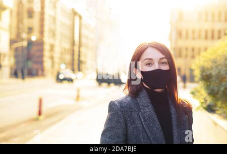 Rotschopf kaukasische Frau auf einer hellen sonnigen leeren Straße in schwarzer Schutzmaske. Revival nach Coronavirus. Stockfoto