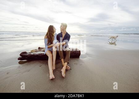 Pärchen, die bei Sonnenuntergang auf einem toten Baumstamm an einem Strand in Bali sitzen und sich anguckte, mit einem Hund im Hintergrund Stockfoto