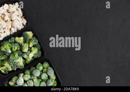 Rahmen von gefrorenem Gemüse in Behältern auf schwarzem Hintergrund. Kohl mischen. Rosenkohl, Brokkoli, Blumenkohl. Leerzeichen für Text. Draufsicht Stockfoto